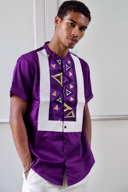 A man wearing a short-sleeved Eloli purple shirt with leather trim on the chest and a round collar, standing in a room with white walls.