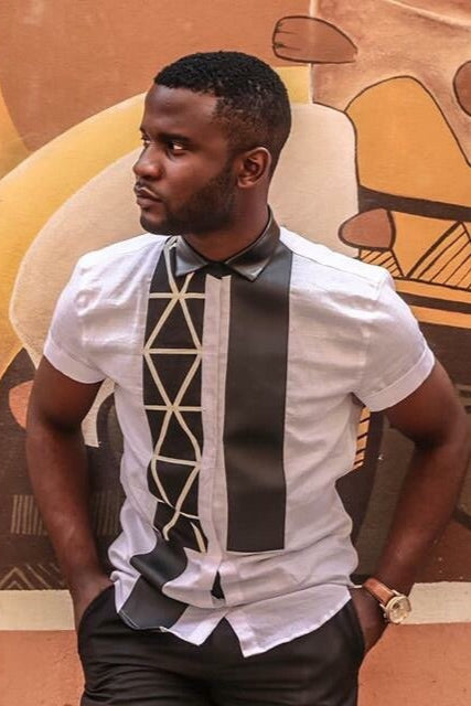 A man wearing a white short-sleeve shirt with a geometric pattern and a black bow tie, standing against a background with abstract art.